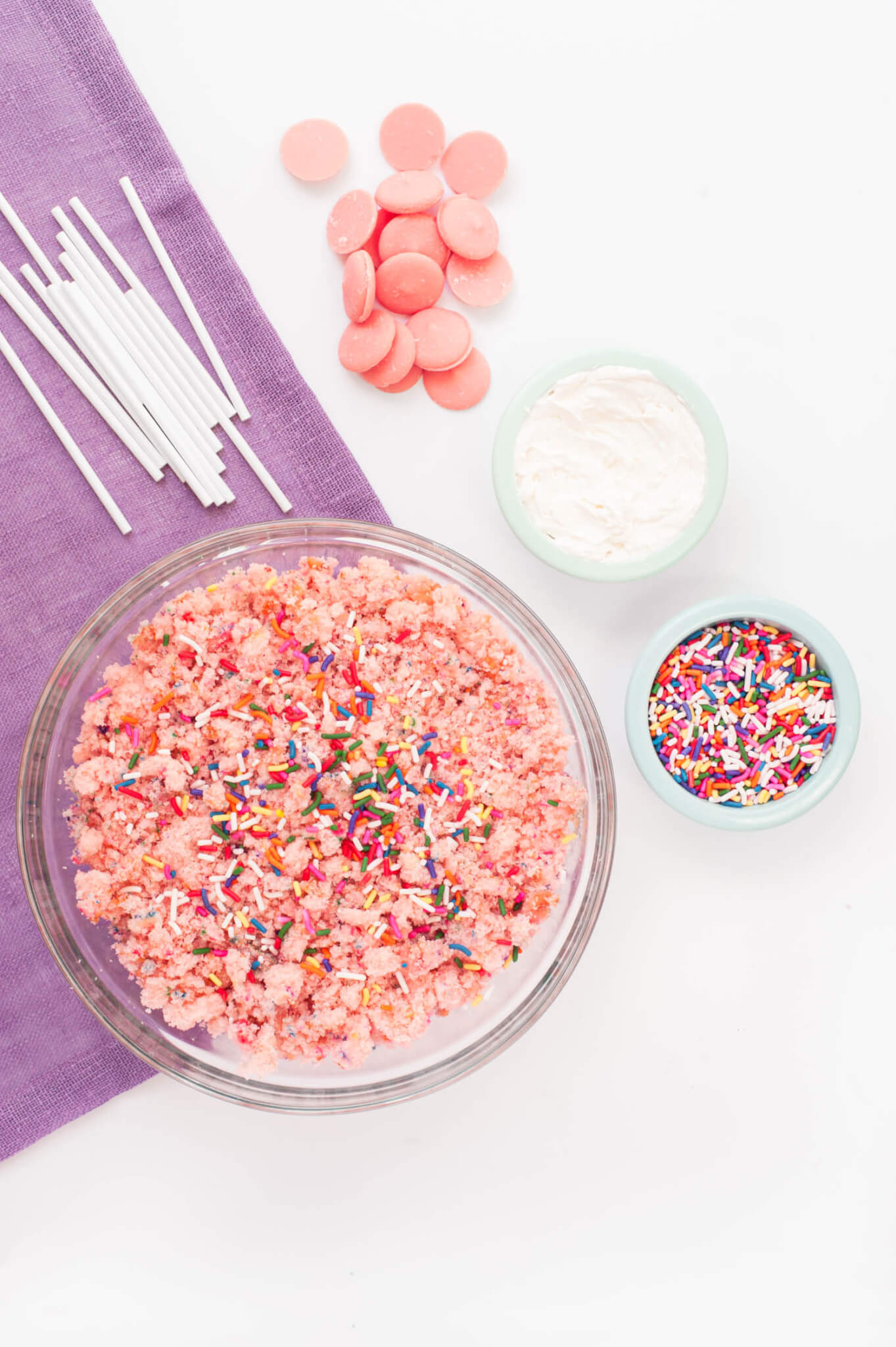 Strawberry Cake Pop Cake crumbles in a glass bowl with cake pop sticks, melting chocolate and sprinkles.