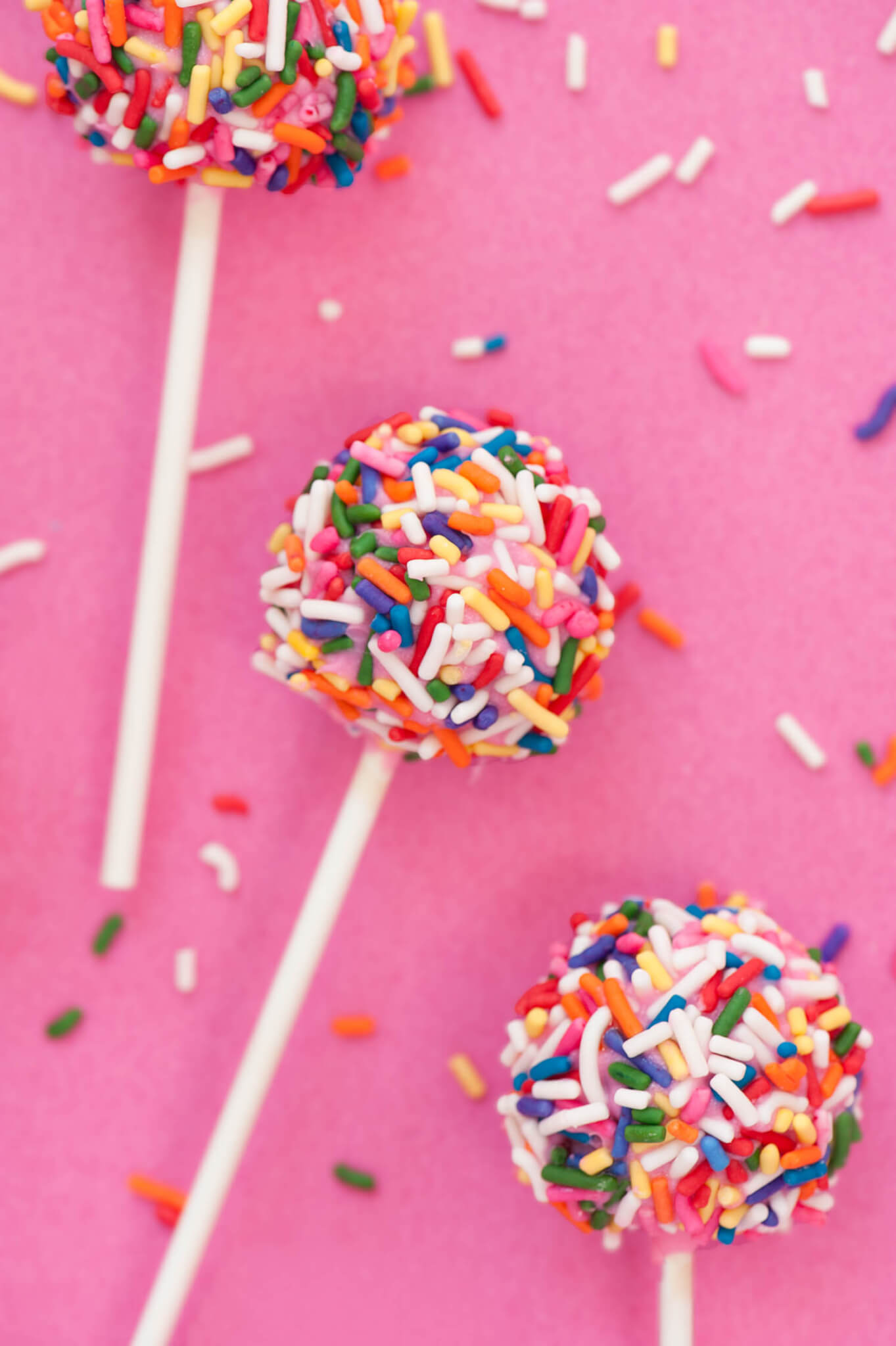 Three cake pops covered in sprinkles displayed on a pink background.